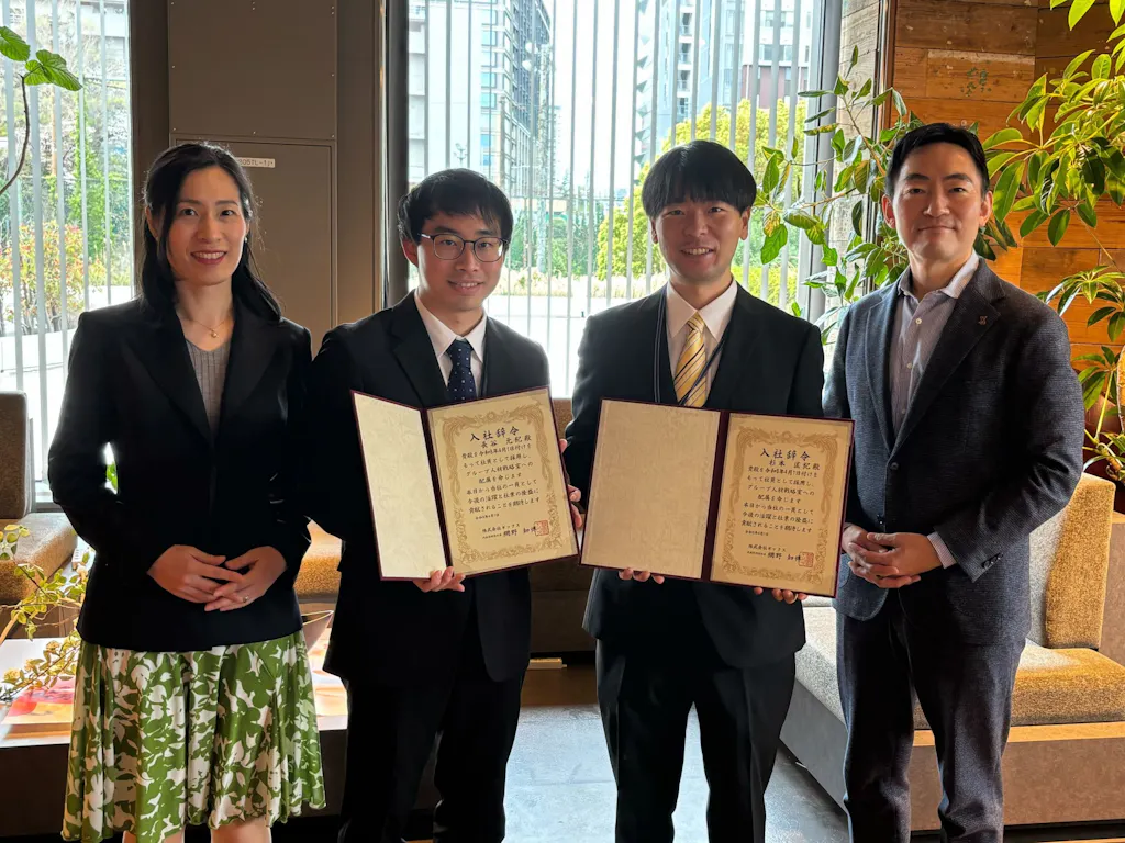 入社式で辞令交付を受けた新入社員2名が、辞令を手に、CEOと人事担当取締役と並んで記念撮影をしている様子